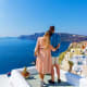 Couple standing on white terrace overlooking Santorini's blue Aegean Sea, caldera cliffs, and whitewashed village buildings